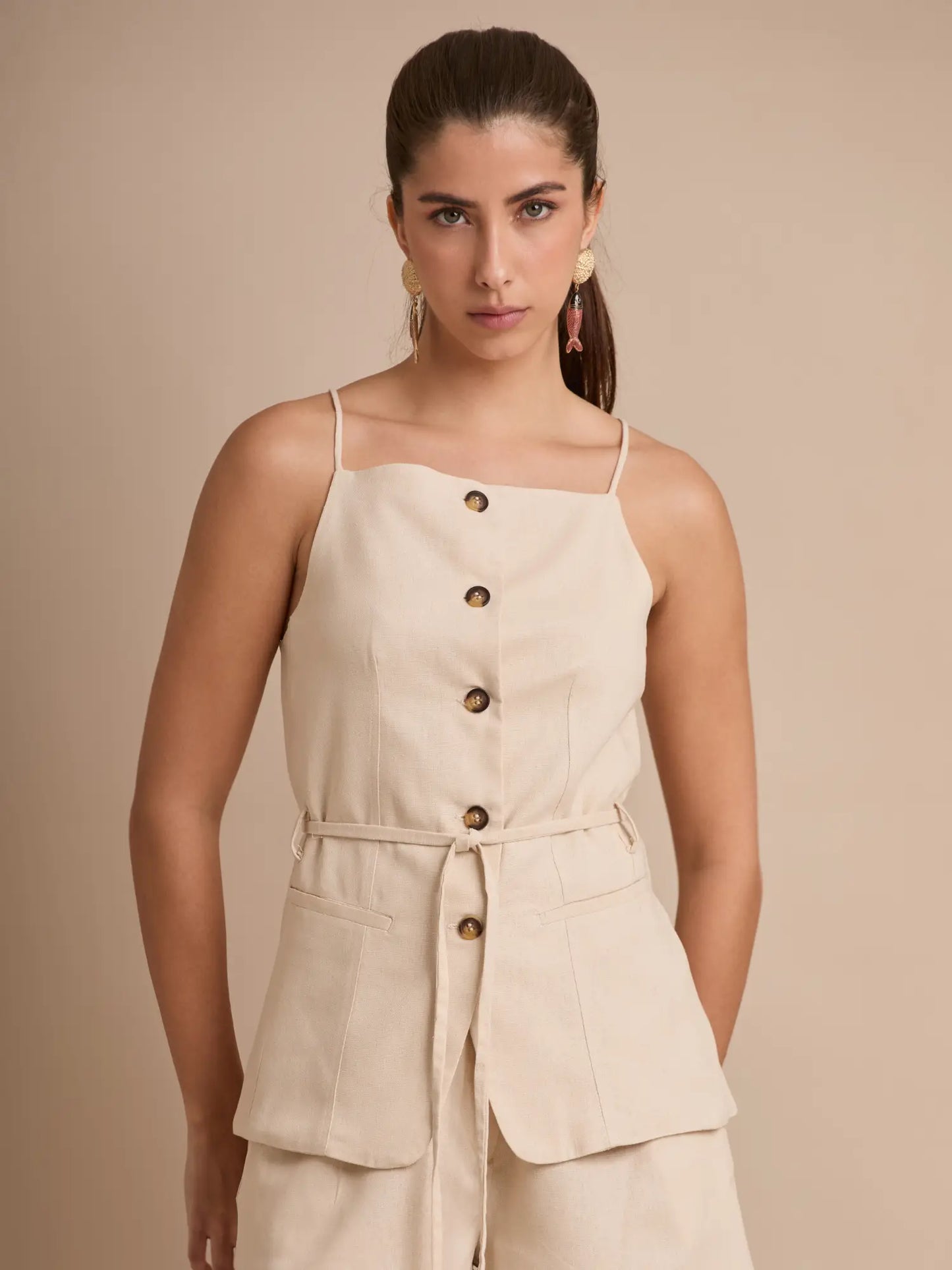 La blusa beige minimalista ofrece líneas limpias, cuello recto y botonadura central. El cinturón del mismo tejido marca la cintura y los bolsillos frontales aportan un aire desenfadado y funcional.