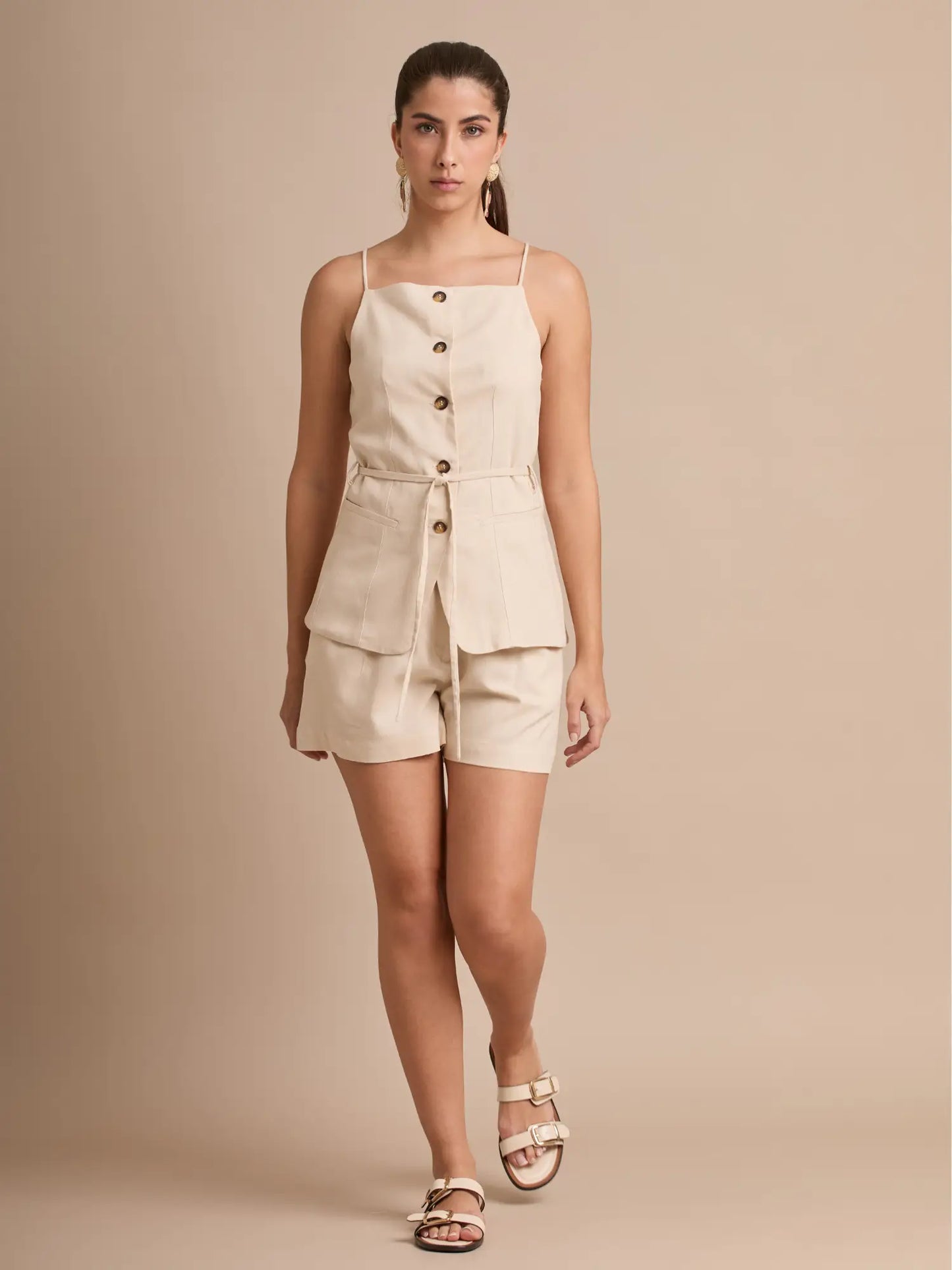 La blusa beige de tirantes tiene escote recto, botones frontales y un cinturón del mismo tejido que marca la cintura. Sus bolsillos tipo cargo al frente le dan un estilo utilitario moderno, ideal para looks de verano cómodos y elegantes.