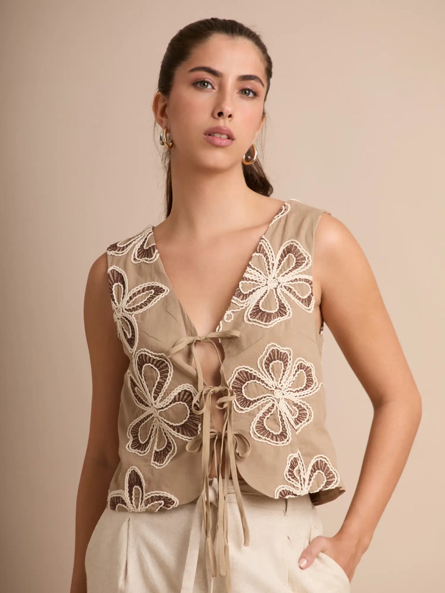 Este chaleco beige bordado ofrece un diseño boho-chic con flores en relieve y amarre frontal, ideal para elevar outfits casuales con un toque artístico y natural.