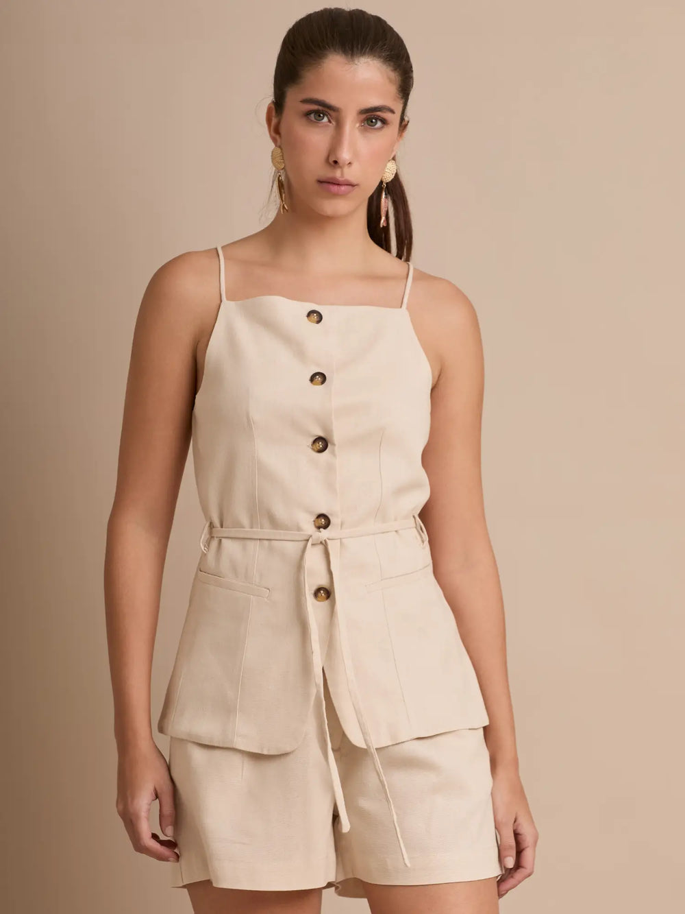 El top beige tiene tiritas finas, corte recto y una hilera de botones que estiliza el torso. Sus bolsillos cargo y el cinturón a juego le dan un toque utilitario y moderno, ideal para combinar con shorts o pantalones amplios.