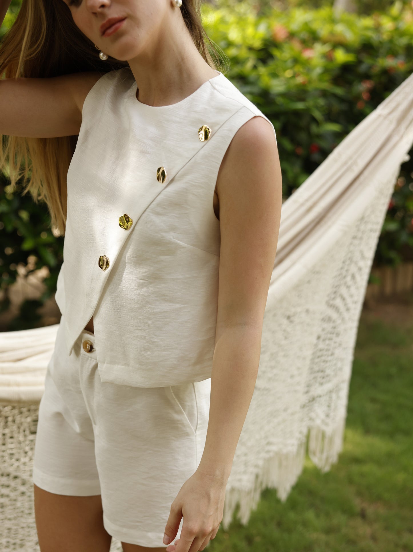 Esta blusa sin mangas tiene un diseño cruzado al frente, asegurado con botones decorativos metálicos dispuestos en diagonal. Presenta un escote redondo y un corte recto que cae de forma relajada sobre la cintura. El bajo es asimétrico, con una ligera abertura en la parte inferior del cruce. Está confeccionada en una tela ligera con textura sutil, ideal para un look fresco y elegante en climas cálidos