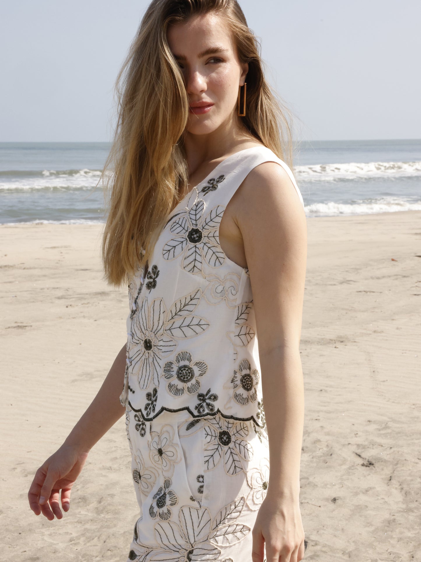 Esta blusa sin mangas tiene un estilo fresco y bohemio, ideal para climas cálidos. Su diseño presenta un escote en "V" y una abertura frontal con botones forrados que le dan un toque artesanal. Está confeccionada en una tela ligera y decorada con bordados florales de gran tamaño, a juego con el short. El bajo tiene un acabado festoneado que sigue el contorno de los bordados, aportando un detalle femenino y delicado. El corte es recto y ligeramente suelto, lo que proporciona comodidad sin perder elegancia. P