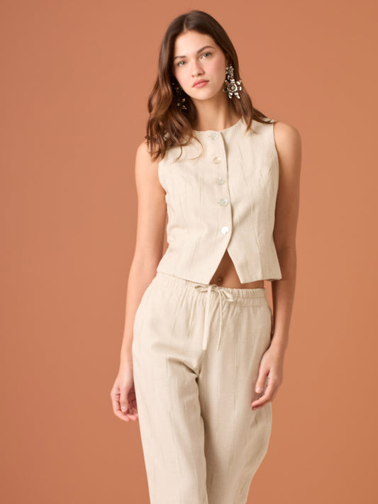 Blusa sin mangas de corte limpio y femenino. Cuello redondo y botonadura frontal con botones tipo joya; pinzas que definen el torso y bajo ligeramente curvado con pequeña abertura al centro. El sutil rayado vertical estiliza la figura. Ideal para combinar con el pantalón a juego o faldas altas para un look pulido.