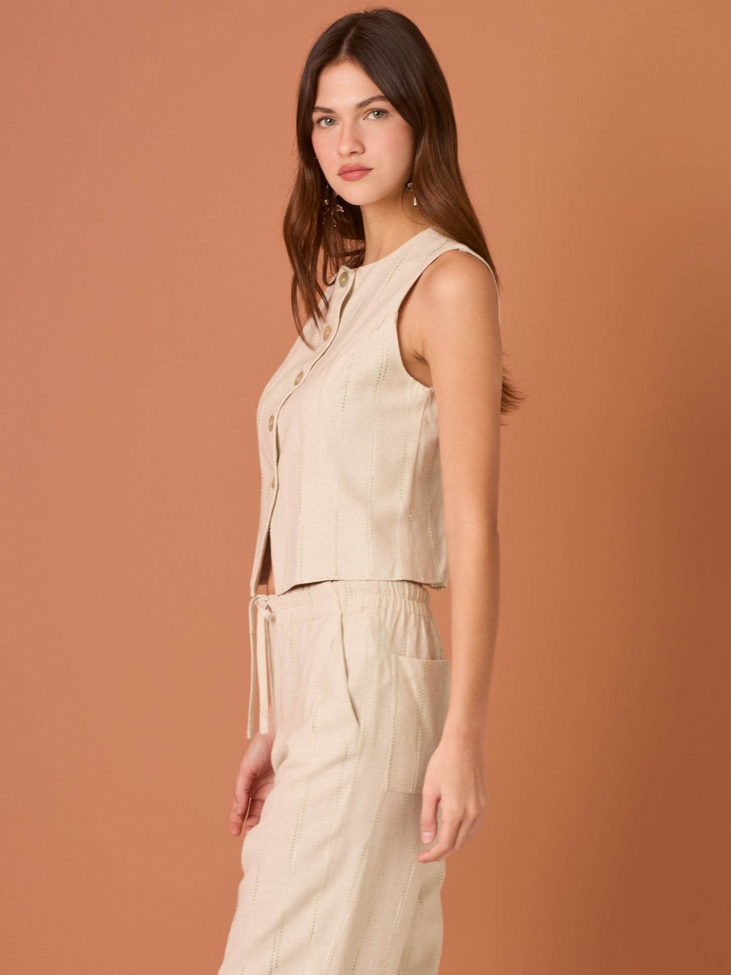 Blusa sin mangas de corte limpio y femenino. Cuello redondo y botonadura frontal con botones tipo joya; pinzas que definen el torso y bajo ligeramente curvado con pequeña abertura al centro. El sutil rayado vertical estiliza la figura. Ideal para combinar con el pantalón a juego o faldas altas para un look pulido.