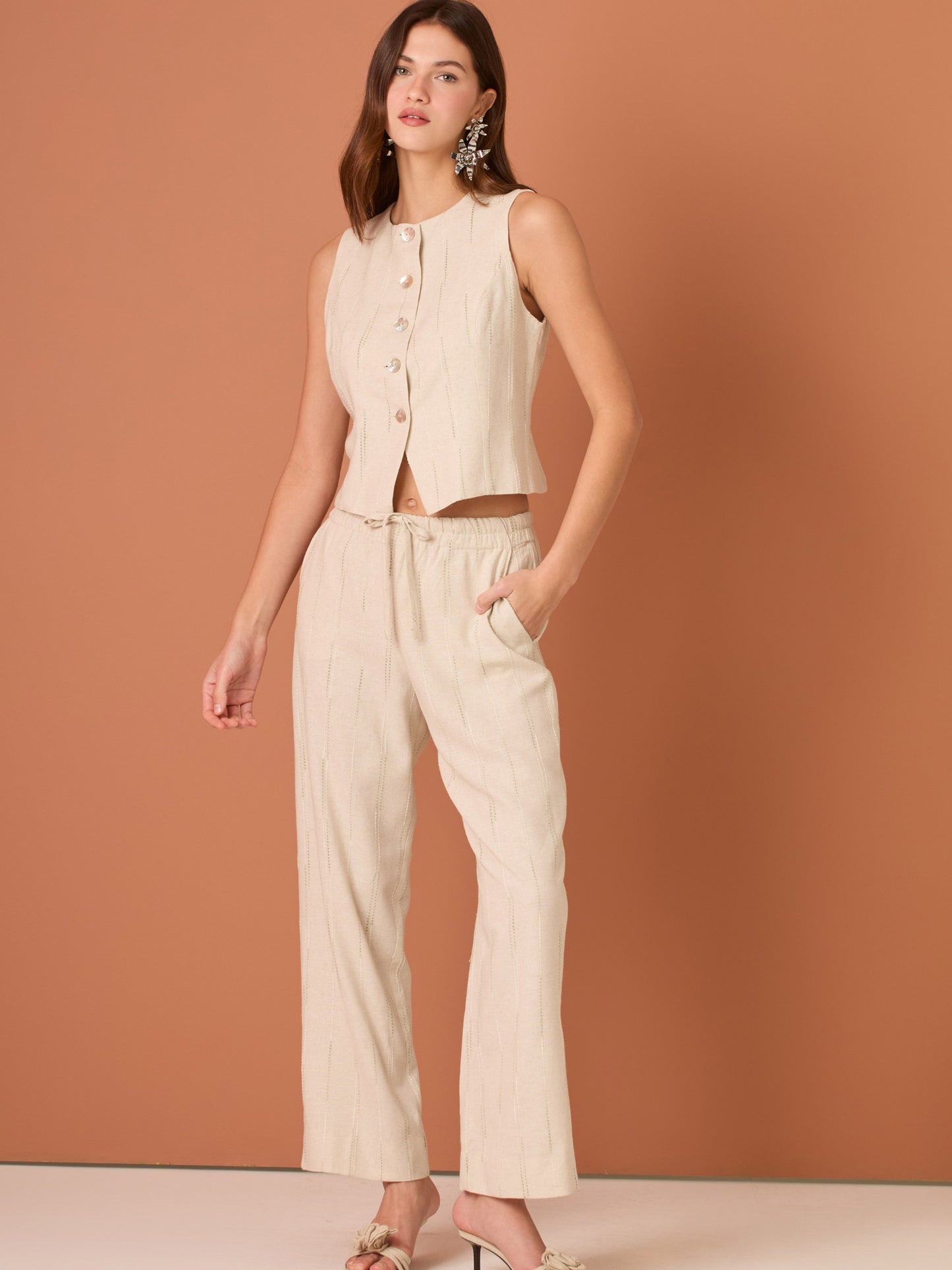 Blusa sin mangas de corte limpio y femenino. Cuello redondo y botonadura frontal con botones tipo joya; pinzas que definen el torso y bajo ligeramente curvado con pequeña abertura al centro. El sutil rayado vertical estiliza la figura. Ideal para combinar con el pantalón a juego o faldas altas para un look pulido.