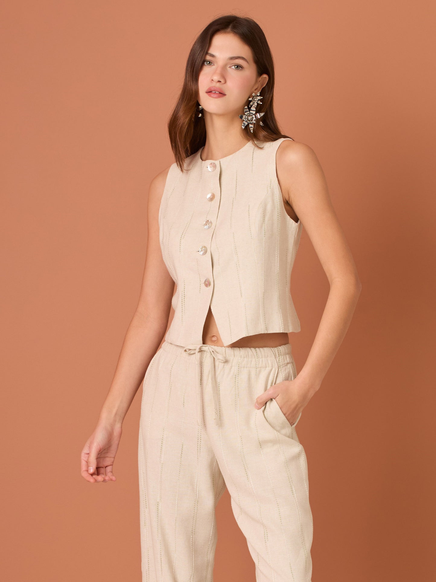 Blusa sin mangas de corte limpio y femenino. Cuello redondo y botonadura frontal con botones tipo joya; pinzas que definen el torso y bajo ligeramente curvado con pequeña abertura al centro. El sutil rayado vertical estiliza la figura. Ideal para combinar con el pantalón a juego o faldas altas para un look pulido.