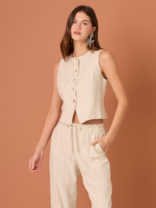 Blusa sin mangas de corte limpio y femenino. Cuello redondo y botonadura frontal con botones tipo joya; pinzas que definen el torso y bajo ligeramente curvado con pequeña abertura al centro. El sutil rayado vertical estiliza la figura. Ideal para combinar con el pantalón a juego o faldas altas para un look pulido.