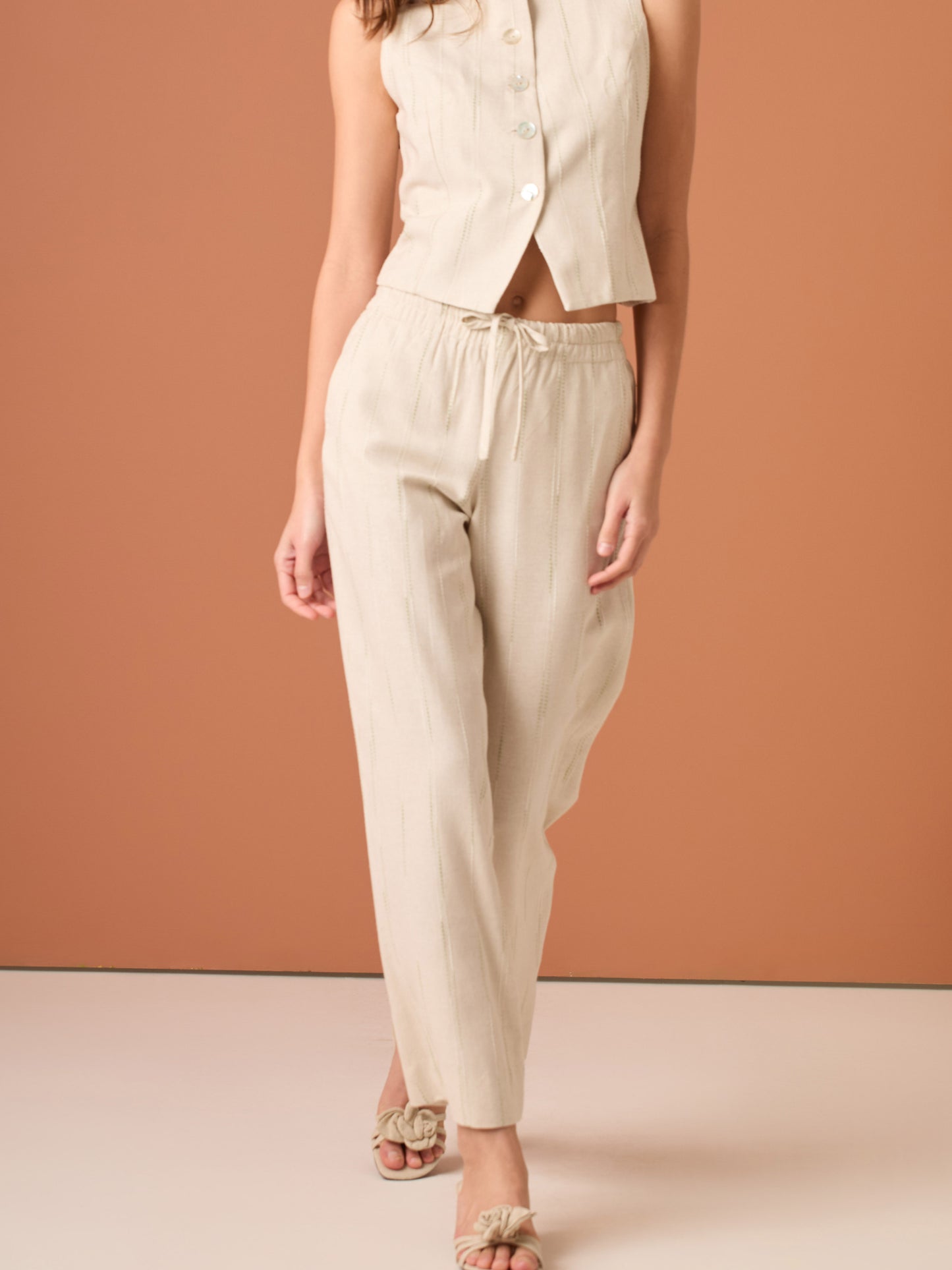 Blusa sin mangas de corte limpio y femenino. Cuello redondo y botonadura frontal con botones tipo joya; pinzas que definen el torso y bajo ligeramente curvado con pequeña abertura al centro. El sutil rayado vertical estiliza la figura. Ideal para combinar con el pantalón a juego o faldas altas para un look pulido.