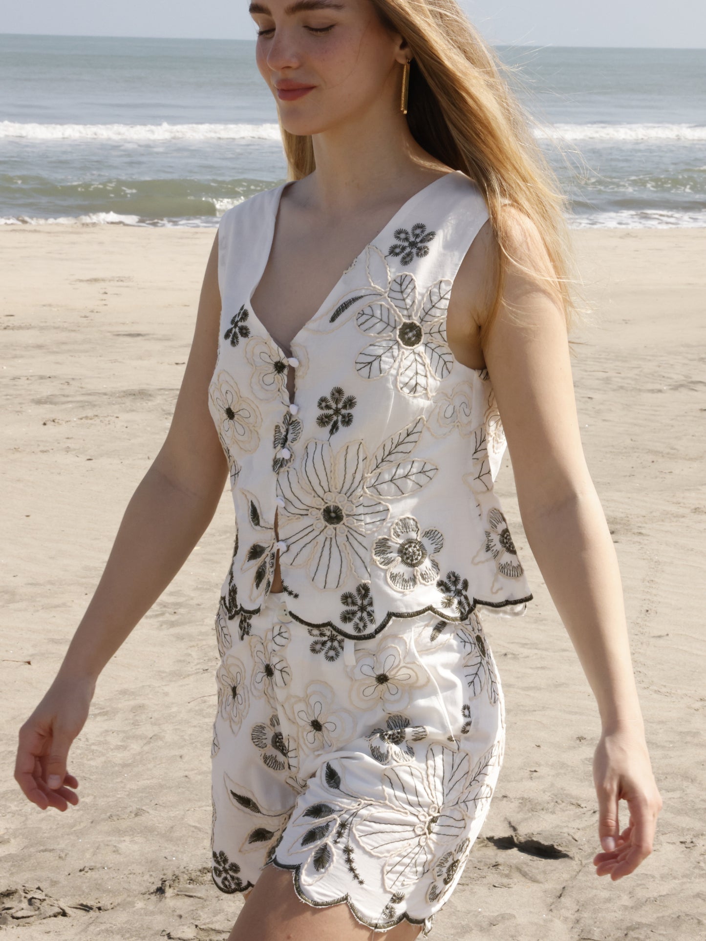 Este short presenta un diseño fresco y veraniego, confeccionado en una tela ligera con bordados florales de gran detalle. Tiene pretina con trabillas para cinturón, cierre frontal con botón y cremallera, y bolsillos laterales. El dobladillo inferior tiene un acabado festoneado que sigue el contorno de los bordados, aportando un toque femenino y artesanal. Es una prenda ideal para un look relajado y elegante de playa o verano