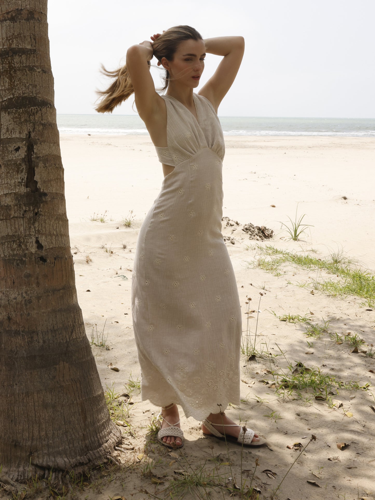 Este vestido es largo, de estilo veraniego y confeccionado en una tela ligera de tono beige claro. Tiene escote halter en V con detalle de bordado calado en todo el tejido, creando pequeños motivos florales que le dan un aire romántico y artesanal. Se ajusta suavemente al cuerpo, marcando la figura sin ser ceñido, y cae recto hasta los tobillos. El diseño es ideal para climas cálidos y ocasiones casuales o de playa, combinando frescura con un toque femenino y elegante