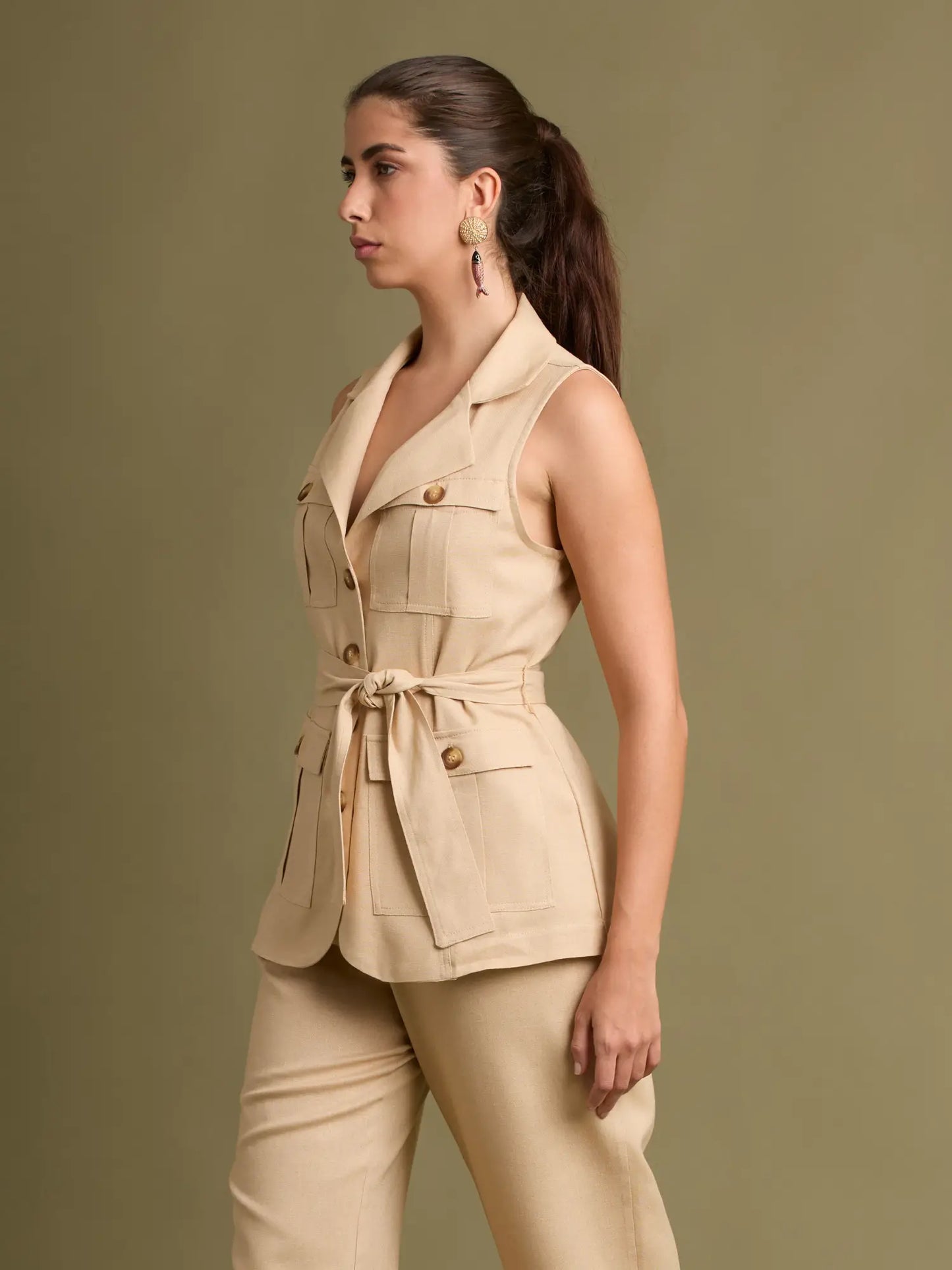 Este chaleco beige utilitario destaca por sus bolsillos de parche, botones en tono madera y un cinturón que define la cintura. Su cuello tipo solapa aporta un toque elegante y moderno.