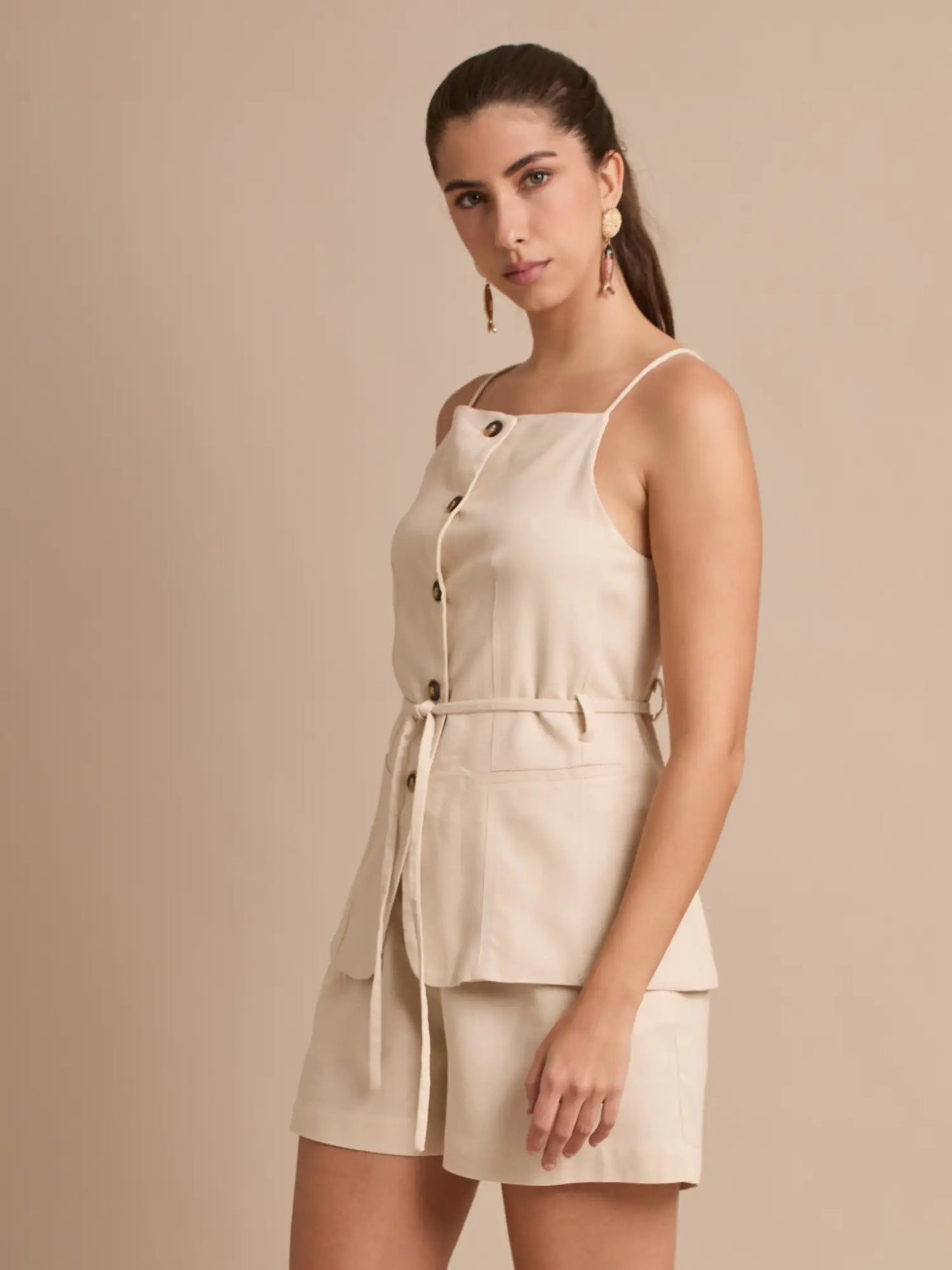 Este top beige tipo chaleco combina tirantes finos con una botonadura central y un cinturón que se ata a la cintura. Los bolsillos frontales alargan la silueta y añaden un aire casual-chic perfecto para el día a día.