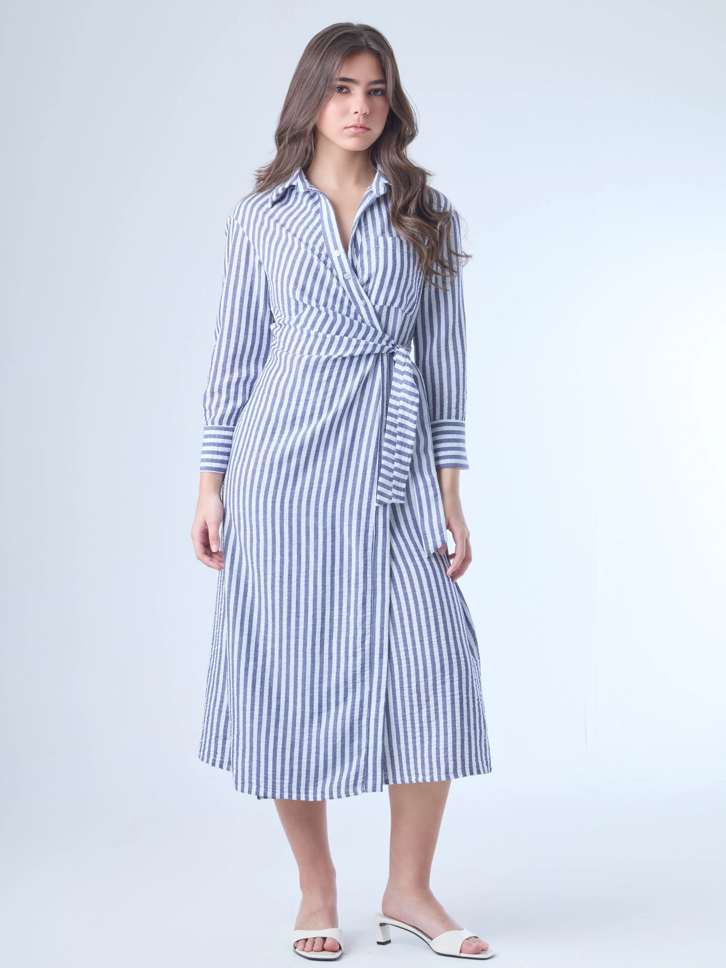 Vestido camisero midi con diseño de rayas verticales. Tiene cuello clásico, mangas largas con puños y un corte cruzado al frente que se ajusta con un lazo a la cintura. La falda cae con movimiento sutil y el ajuste en la cintura define la silueta de forma favorecedora. Es una prenda cómoda, estructurada y con un aire sofisticado.

Detalles:

Color: Azul
