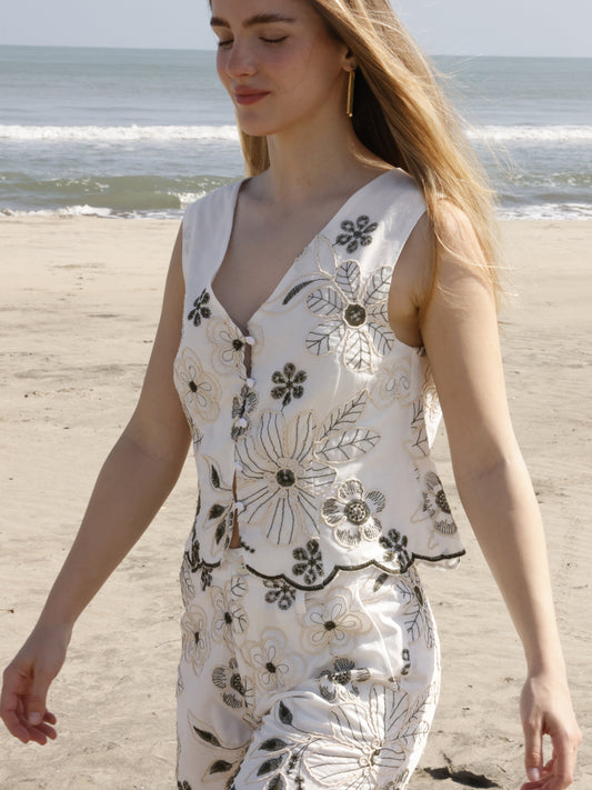 Esta blusa sin mangas tiene un estilo fresco y bohemio, ideal para climas cálidos. Su diseño presenta un escote en "V" y una abertura frontal con botones forrados que le dan un toque artesanal. Está confeccionada en una tela ligera y decorada con bordados florales de gran tamaño, a juego con el short. El bajo tiene un acabado festoneado que sigue el contorno de los bordados, aportando un detalle femenino y delicado. El corte es recto y ligeramente suelto, lo que proporciona comodidad sin perder elegancia. P