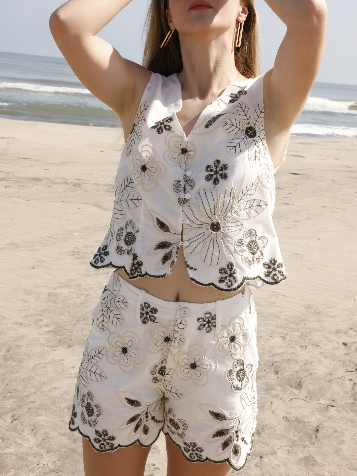 Este short presenta un diseño fresco y veraniego, confeccionado en una tela ligera con bordados florales de gran detalle. Tiene pretina con trabillas para cinturón, cierre frontal con botón y cremallera, y bolsillos laterales. El dobladillo inferior tiene un acabado festoneado que sigue el contorno de los bordados, aportando un toque femenino y artesanal. Es una prenda ideal para un look relajado y elegante de playa o verano