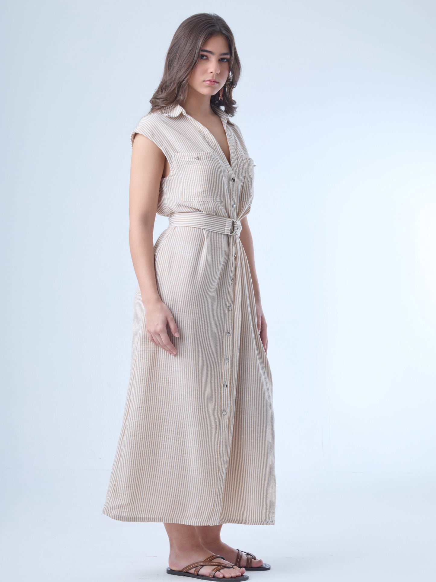 Vestido midi de corte camisero, sin mangas y con hombreras ligeras. Presenta escote en "V" profundo, bolsillos de tapa en el pecho y botonadura frontal completa. Incluye un cinturón del mismo tejido con hebilla doble para acentuar la cintura, y una abertura en la parte inferior que facilita el movimiento. El patrón de rayas verticales estiliza la silueta y aporta un aire fresco y casual.

Detalles:

Color: Sand

Material: 100% Poliéster&nbsp;