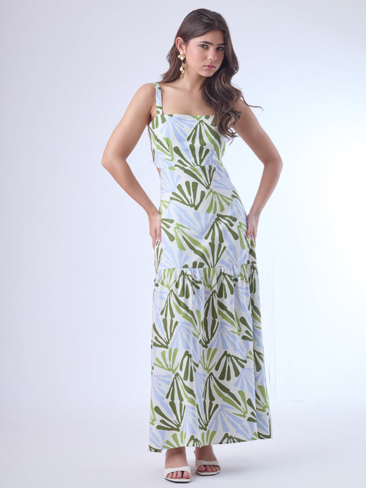 Este vestido largo presenta un diseño fresco y moderno con estampado botánico en tonos verde oliva, verde lima y azul claro sobre fondo blanco. Tiene tirantes anchos y escote recto, que aportan estructura y realzan los hombros. Los laterales descubiertos le dan un toque contemporáneo y femenino, acentuando la figura de forma sutil. La falda cae en línea recta con un corte bajo y un ligero fruncido, lo que aporta movimiento y una silueta relajada. Es una prenda ideal para climas cálidos y ocasiones casuales 