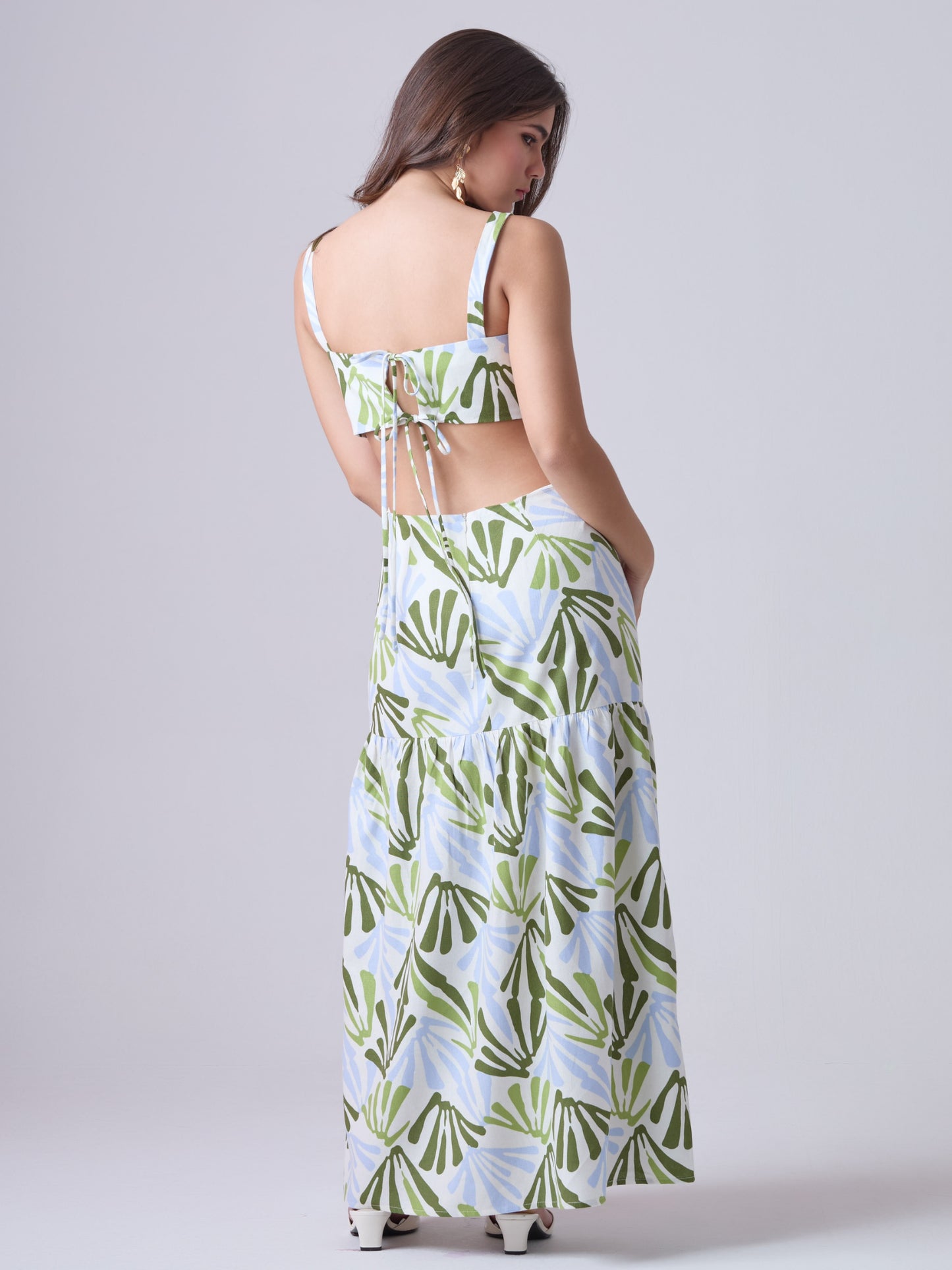 Este vestido largo presenta un diseño fresco y moderno con estampado botánico en tonos verde oliva, verde lima y azul claro sobre fondo blanco. Tiene tirantes anchos y escote recto, que aportan estructura y realzan los hombros. Los laterales descubiertos le dan un toque contemporáneo y femenino, acentuando la figura de forma sutil. La falda cae en línea recta con un corte bajo y un ligero fruncido, lo que aporta movimiento y una silueta relajada. Es una prenda ideal para climas cálidos y ocasiones casuales 