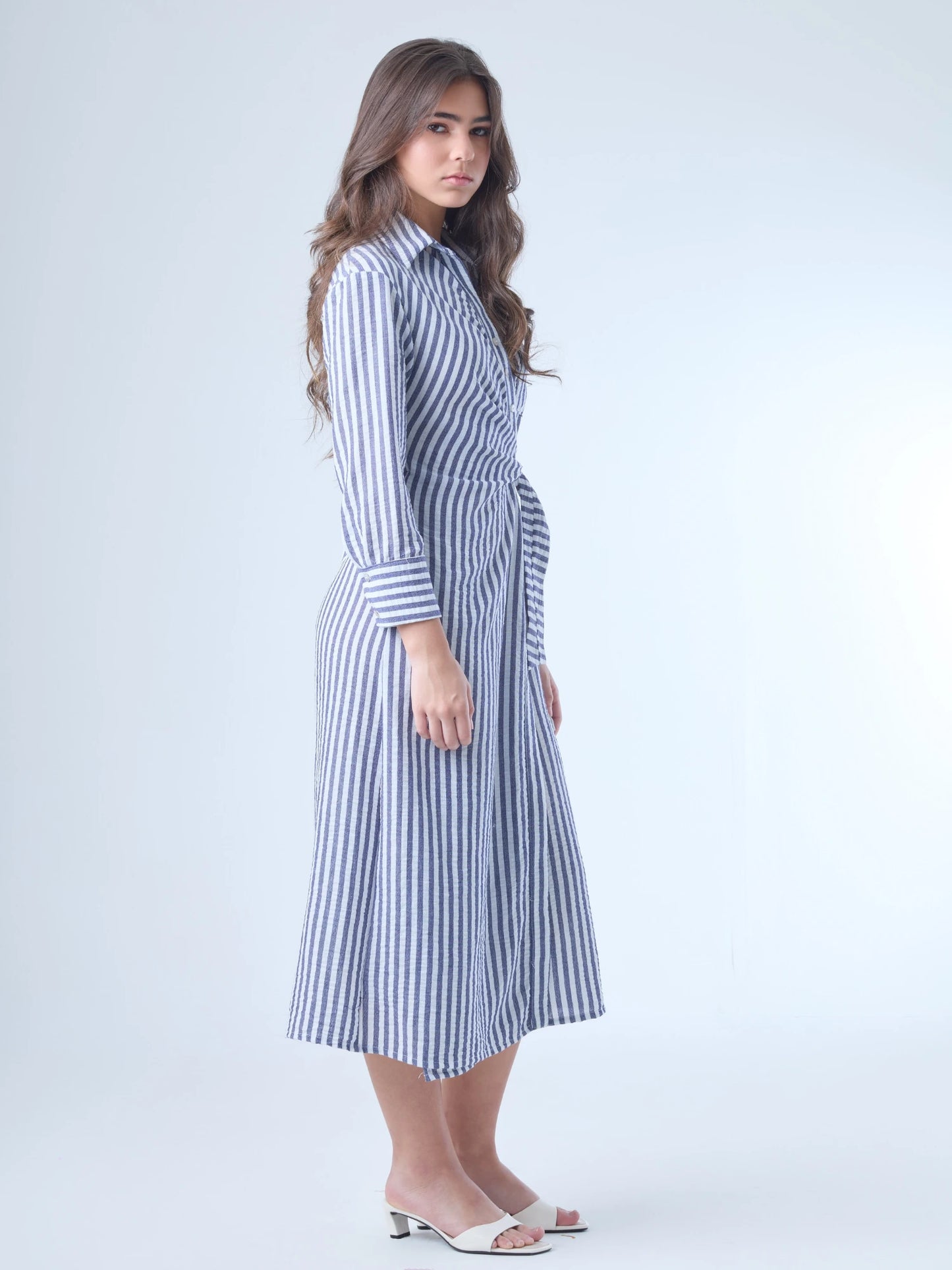 Vestido camisero midi con diseño de rayas verticales. Tiene cuello clásico, mangas largas con puños y un corte cruzado al frente que se ajusta con un lazo a la cintura. La falda cae con movimiento sutil y el ajuste en la cintura define la silueta de forma favorecedora. Es una prenda cómoda, estructurada y con un aire sofisticado.

Detalles:

Color: Azul