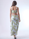 Este vestido largo presenta un diseño fresco y moderno con estampado botánico en tonos verde oliva, verde lima y azul claro sobre fondo blanco. Tiene tirantes anchos y escote recto, que aportan estructura y realzan los hombros. Los laterales descubiertos le dan un toque contemporáneo y femenino, acentuando la figura de forma sutil. La falda cae en línea recta con un corte bajo y un ligero fruncido, lo que aporta movimiento y una silueta relajada. Es una prenda ideal para climas cálidos y ocasiones casuales 