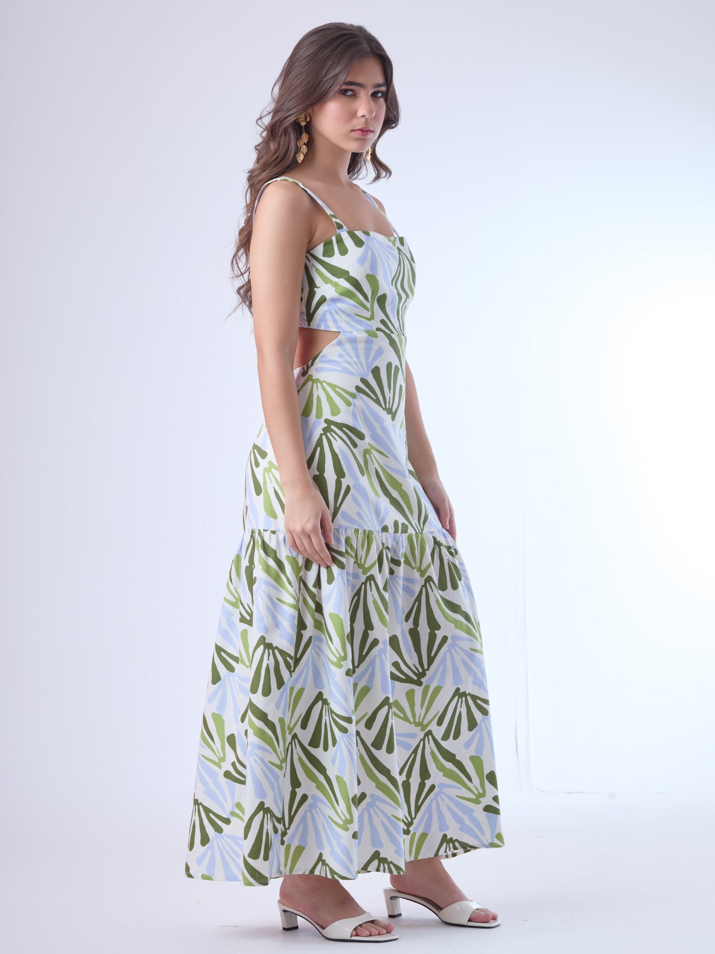 Este vestido largo presenta un diseño fresco y moderno con estampado botánico en tonos verde oliva, verde lima y azul claro sobre fondo blanco. Tiene tirantes anchos y escote recto, que aportan estructura y realzan los hombros. Los laterales descubiertos le dan un toque contemporáneo y femenino, acentuando la figura de forma sutil. La falda cae en línea recta con un corte bajo y un ligero fruncido, lo que aporta movimiento y una silueta relajada. Es una prenda ideal para climas cálidos y ocasiones casuales 