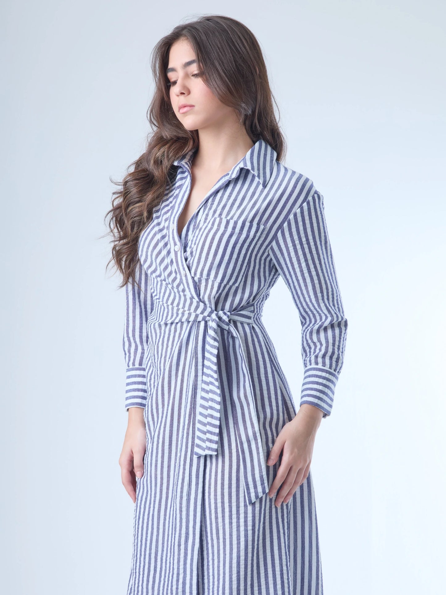Vestido camisero midi con diseño de rayas verticales. Tiene cuello clásico, mangas largas con puños y un corte cruzado al frente que se ajusta con un lazo a la cintura. La falda cae con movimiento sutil y el ajuste en la cintura define la silueta de forma favorecedora. Es una prenda cómoda, estructurada y con un aire sofisticado.

Detalles:

Color: Azul