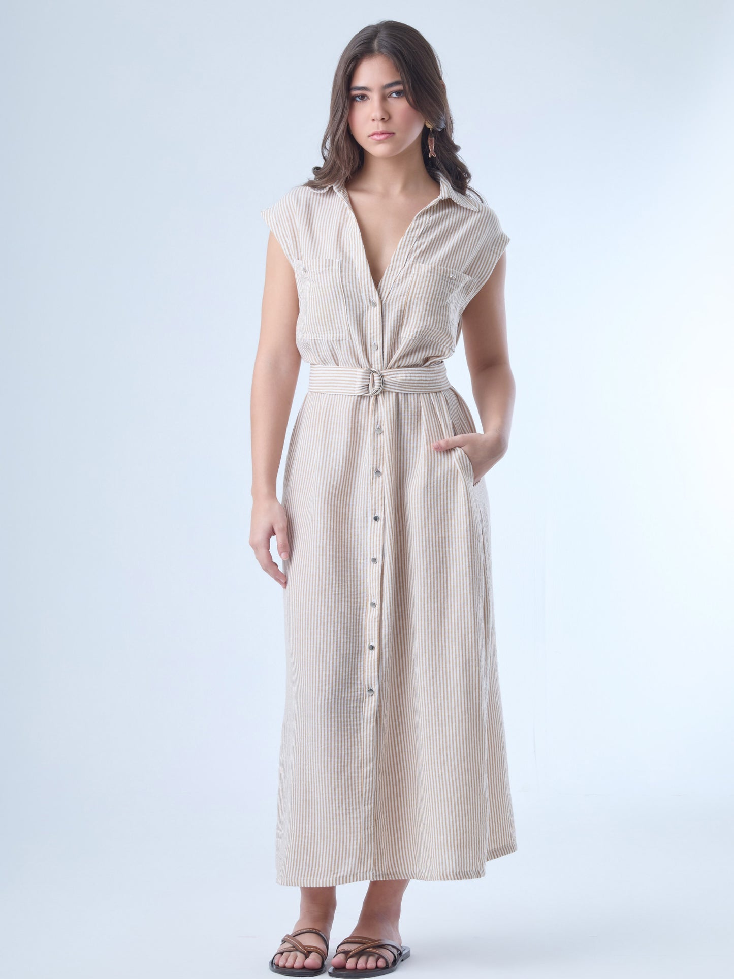 Vestido midi de corte camisero, sin mangas y con hombreras ligeras. Presenta escote en "V" profundo, bolsillos de tapa en el pecho y botonadura frontal completa. Incluye un cinturón del mismo tejido con hebilla doble para acentuar la cintura, y una abertura en la parte inferior que facilita el movimiento. El patrón de rayas verticales estiliza la silueta y aporta un aire fresco y casual.

Detalles:

Color: Sand

Material: 100% Poliéster&nbsp;