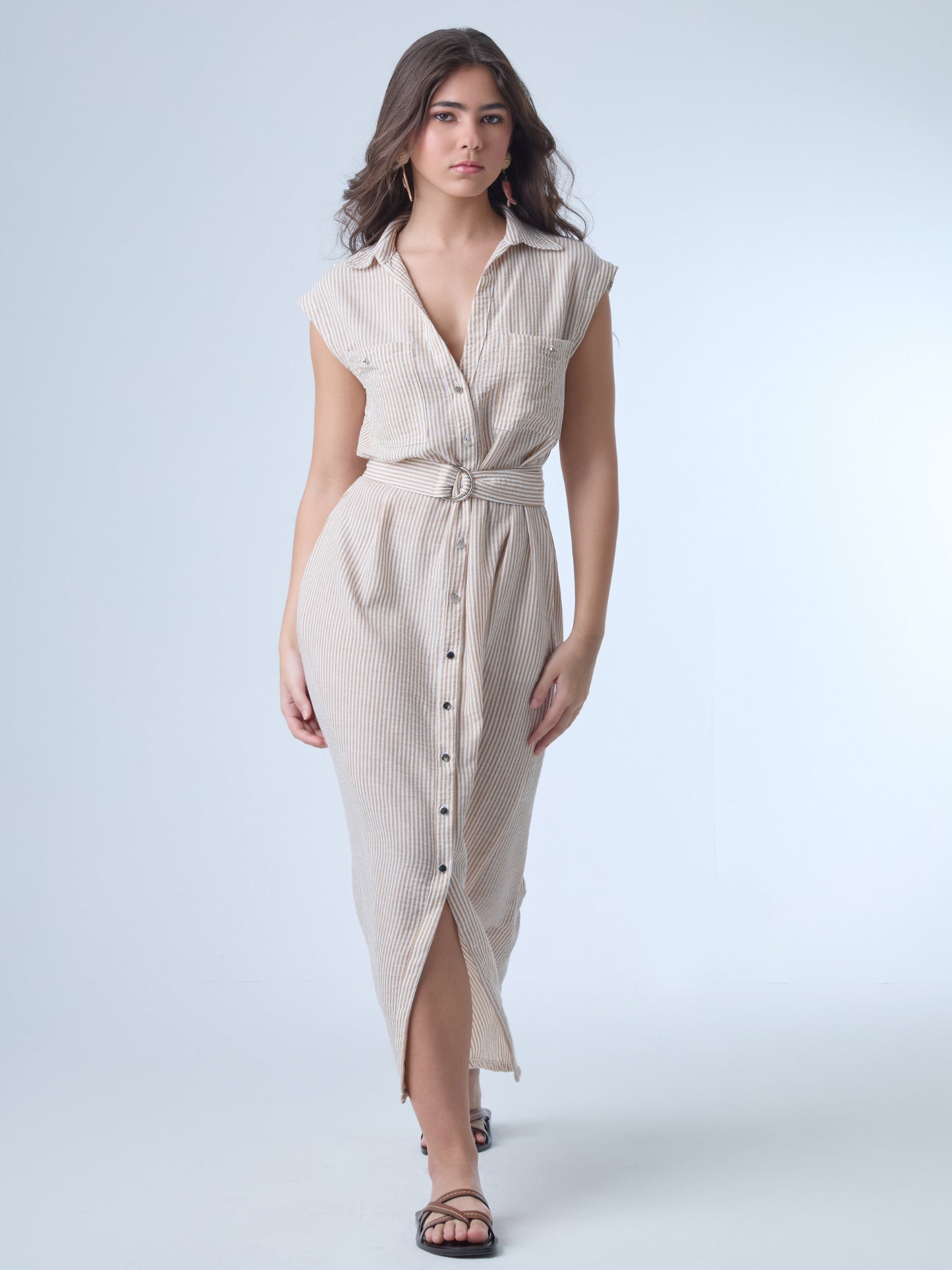 Vestido midi de corte camisero, sin mangas y con hombreras ligeras. Presenta escote en "V" profundo, bolsillos de tapa en el pecho y botonadura frontal completa. Incluye un cinturón del mismo tejido con hebilla doble para acentuar la cintura, y una abertura en la parte inferior que facilita el movimiento. El patrón de rayas verticales estiliza la silueta y aporta un aire fresco y casual.

Detalles:

Color: Sand

Material: 100% Poliéster&nbsp;